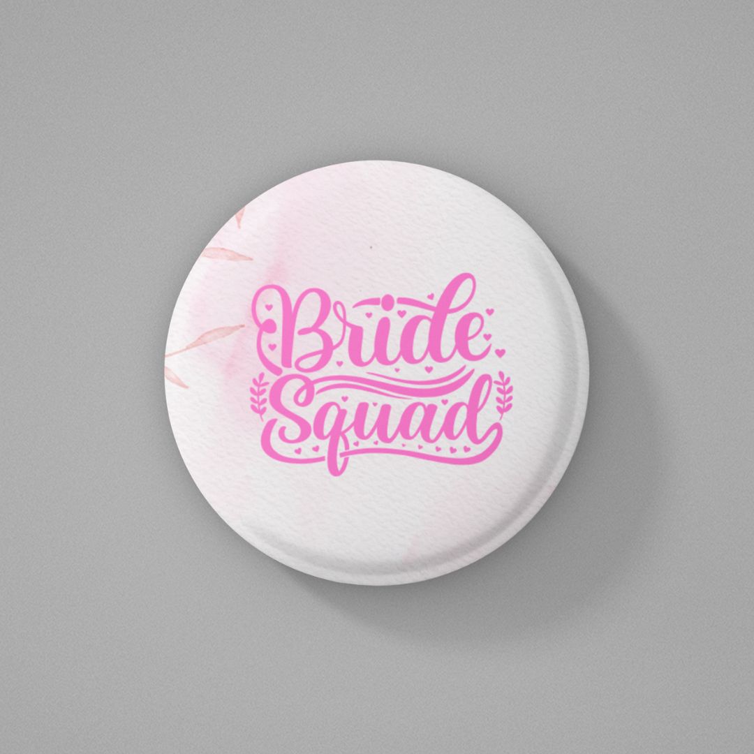 Round white pin badge with pink color 'Bride Squad' text written on it and placed on a gray background