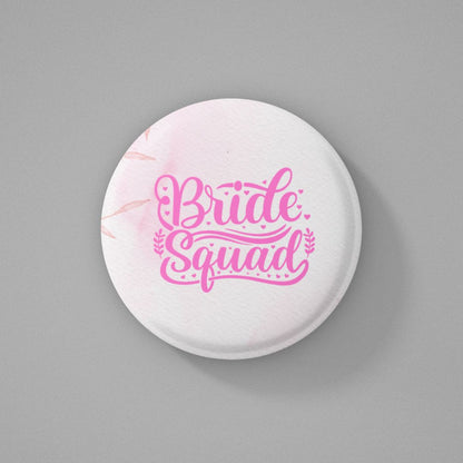 Round white pin badge with pink color 'Bride Squad' text written on it and placed on a gray background
