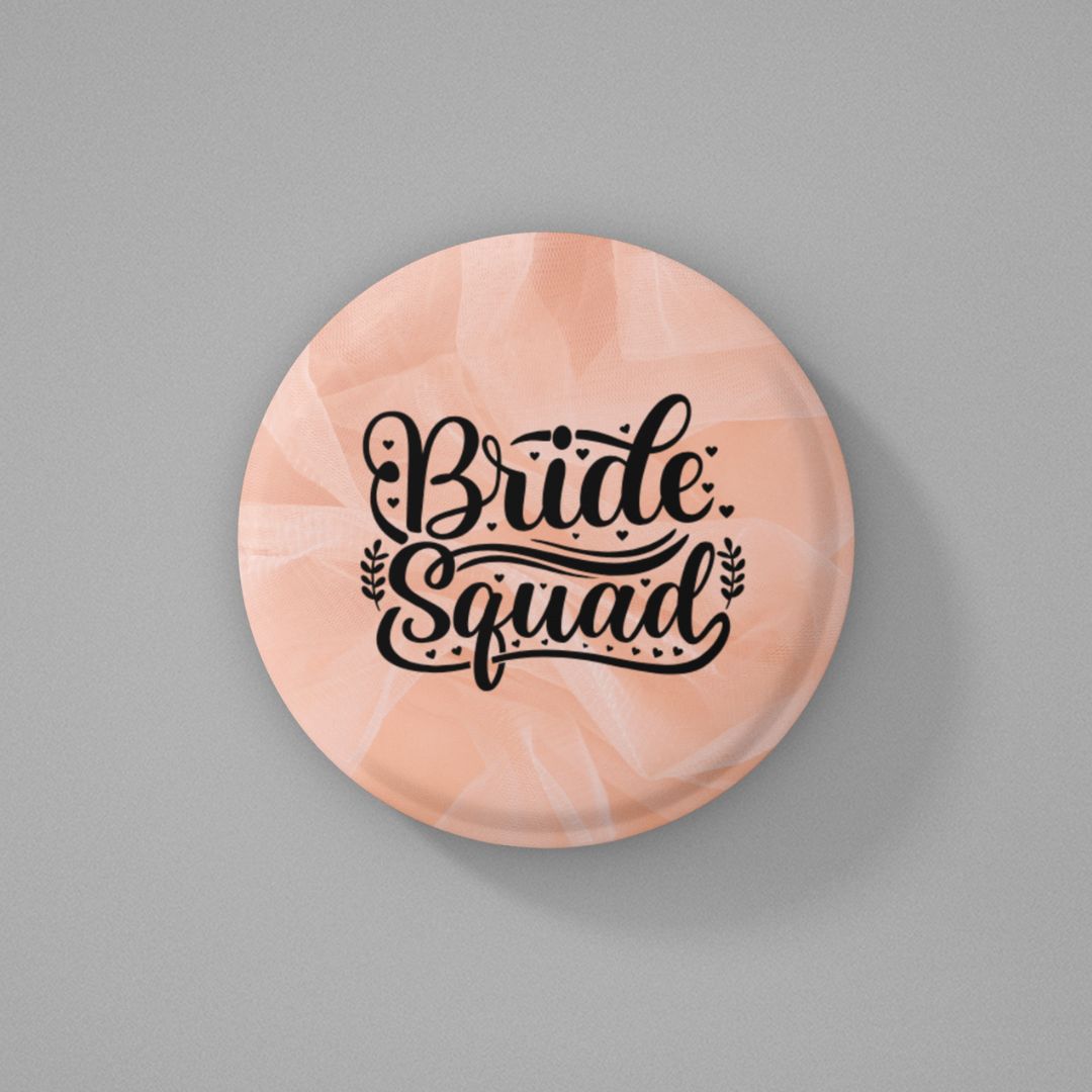 Round pink colored pin badge with 'Bride Squad' text written on it and placed on a gray background