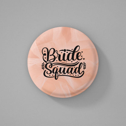 Round pink colored pin badge with 'Bride Squad' text written on it and placed on a gray background